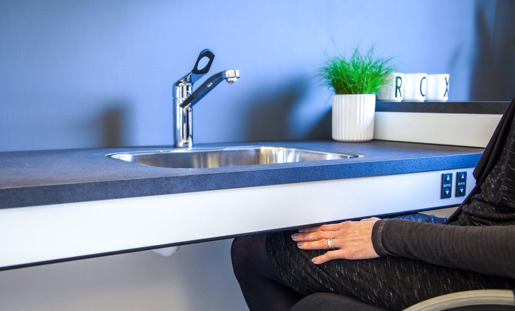 Frau im Rollstuhl an unterfahrbarer, höhenverstellbarer Arbeitsfläche mit flachem Spülenbecken und Armatur mit langem Hebel