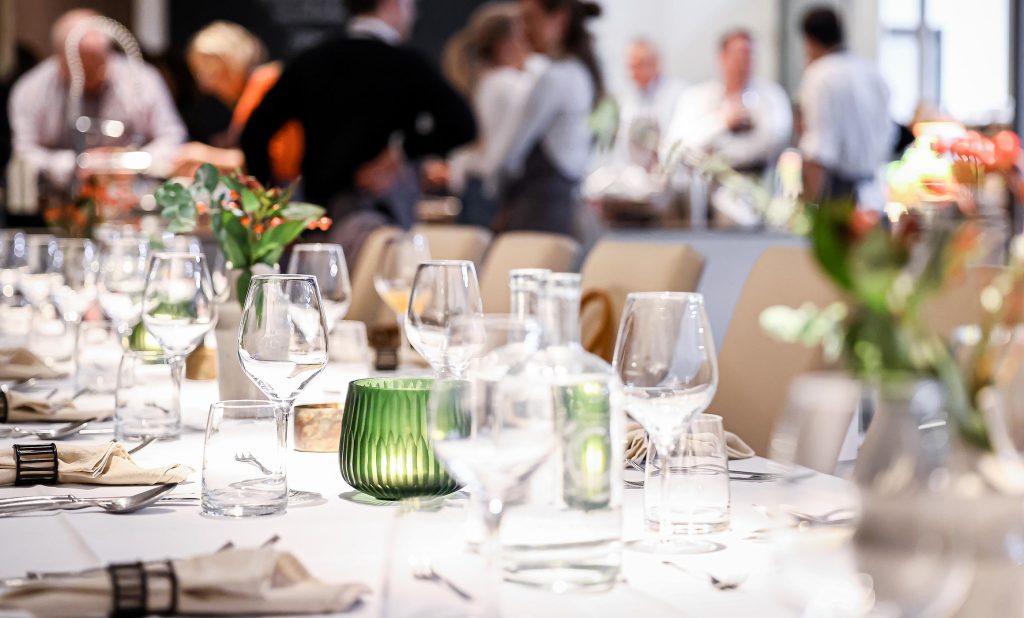 Edel eingedeckte Tafel für ein privates Event in der Cucinaria Kochschule. Gläser, weiße Tischdecke und Personen im Hintergrund.