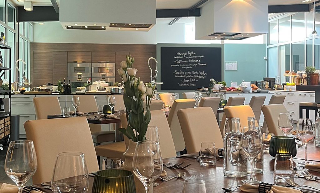 Gedeckte Tafeln in der offenen Kochschulküche von Cucinaria in Hamburg. Durchblick in die Hummel Küchenausstellung.